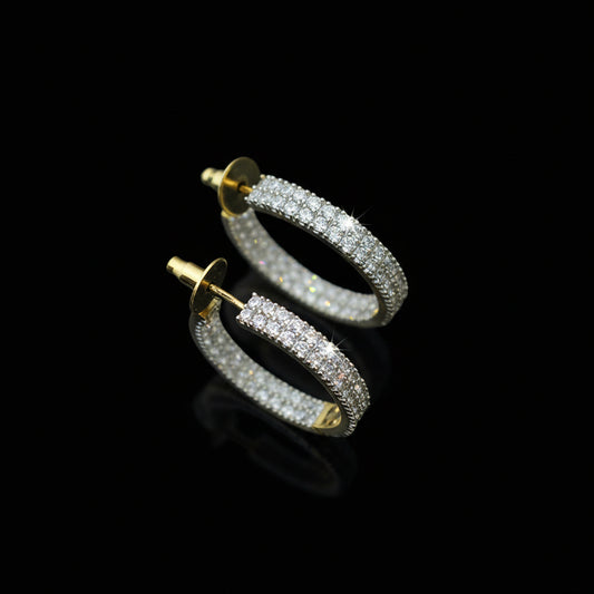 Gold hoop earrings with clear stones on a reflective surface, branded 'SAHELI'.