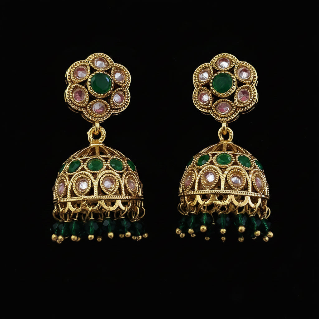 A pair of gold-plated Indian jhumka earrings with green stones and clear rhinestones, displayed on a solid magenta background.