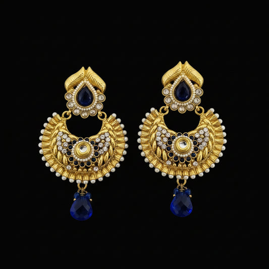 A pair of ornate gold-toned Indian jhumka earrings with dark blue teardrop stones, white faux pearls, and small clear stones, displayed on a dark reflective surface with the brand name 'SAHELI' visible at the top.