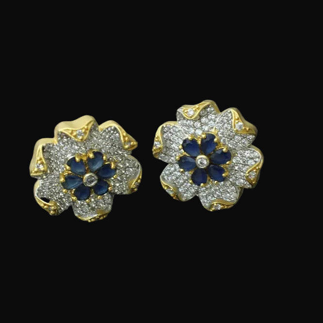 Flower-shaped earrings with blue centers and gold accents on a gray background, featuring the brand 'SAHELI'.