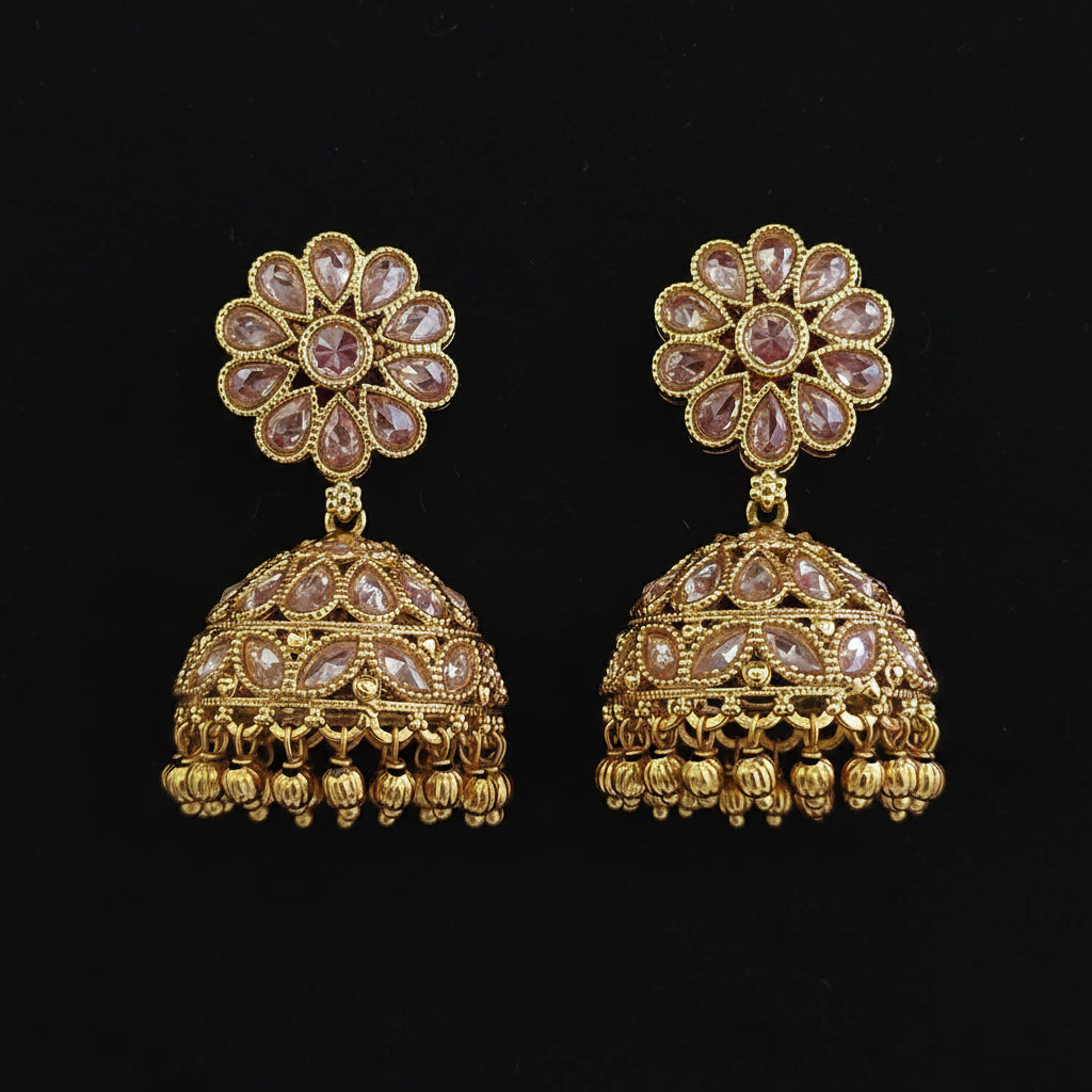 A pair of gold-plated Indian jhumka earrings decorated with clear stones in a floral pattern and a dome-shaped jhumka with hanging bell details, set against a solid dark red background.