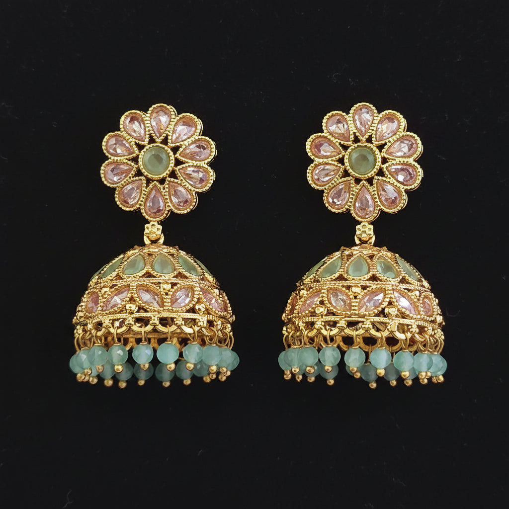 A pair of gold-plated Indian jhumka earrings with light green stone inlays, clear rhinestone flower tops, and dangling light green bead tassels, displayed on a solid dark red background.