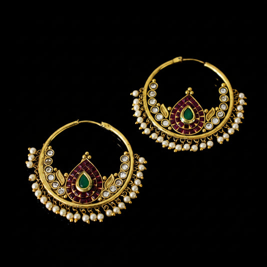 A pair of gold-plated traditional Indian hoop earrings decorated with white pearl-like beads, red enamel, and green stone accents, displayed on a dark reflective surface.