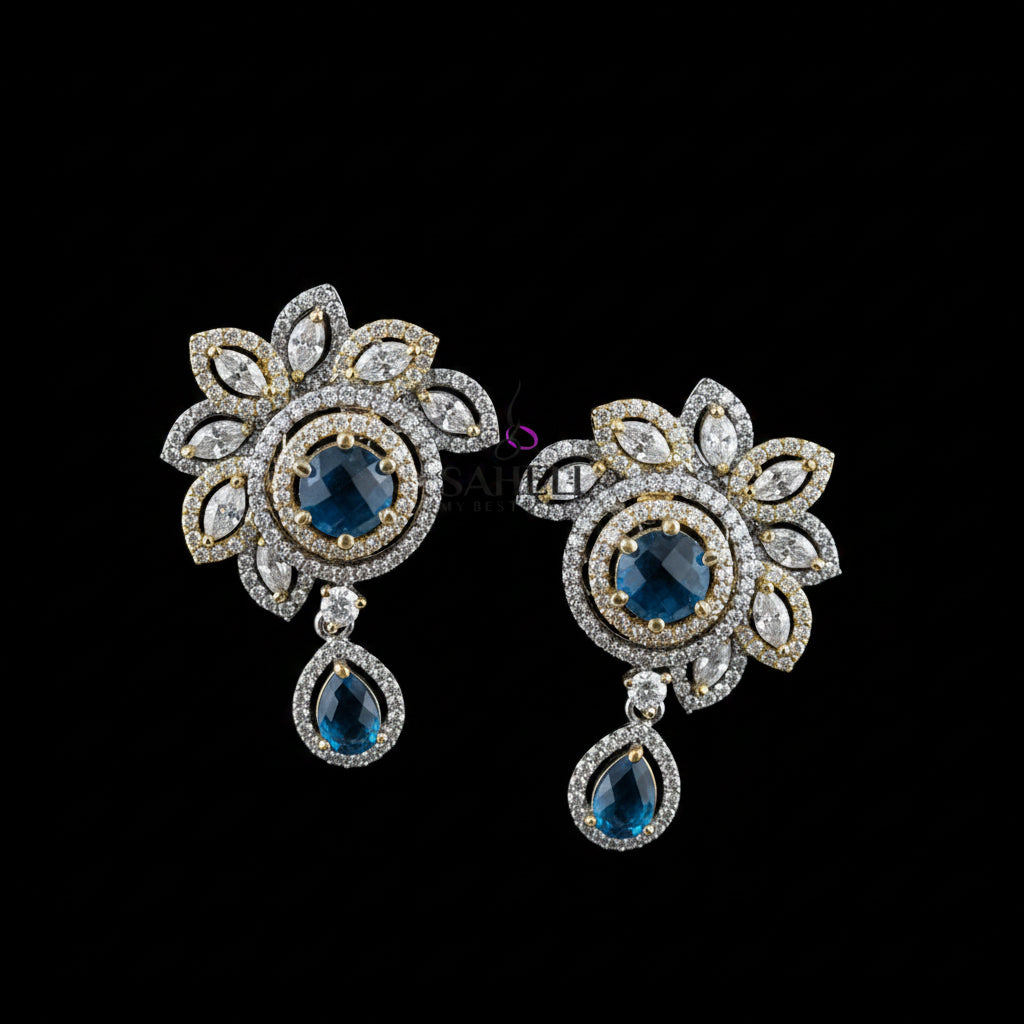 A pair of ornate floral drop earrings displayed on a dark gray stand against a gray background, with the brand name 'SAHELI MY BEST FRIEND' printed above. The earrings feature gold and silver-tone metal settings, clear pavé stones, and deep blue teardrop and round center stones.