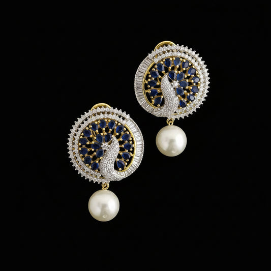 A pair of gold and silver-toned Indian Kundan earrings featuring peacock motifs with deep blue and white stones, finished with a dangling white faux-pearl bead, displayed on a dark reflective surface with the 'SAHELI MY BEST FRIEND' logo in the background.