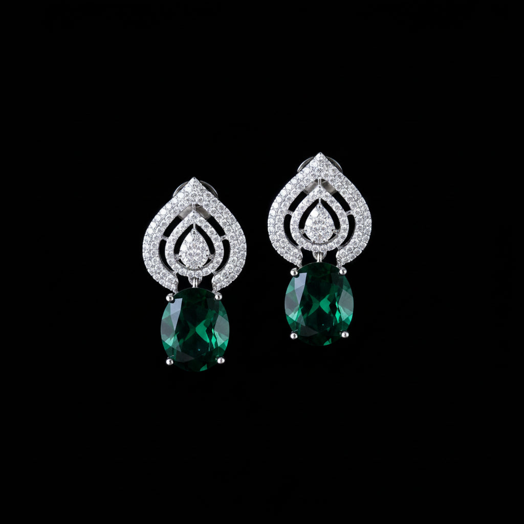 A pair of silver teardrop-shaped earrings with pavé-set clear stones and a round green gemstone dangling below, displayed on a dark reflective surface.