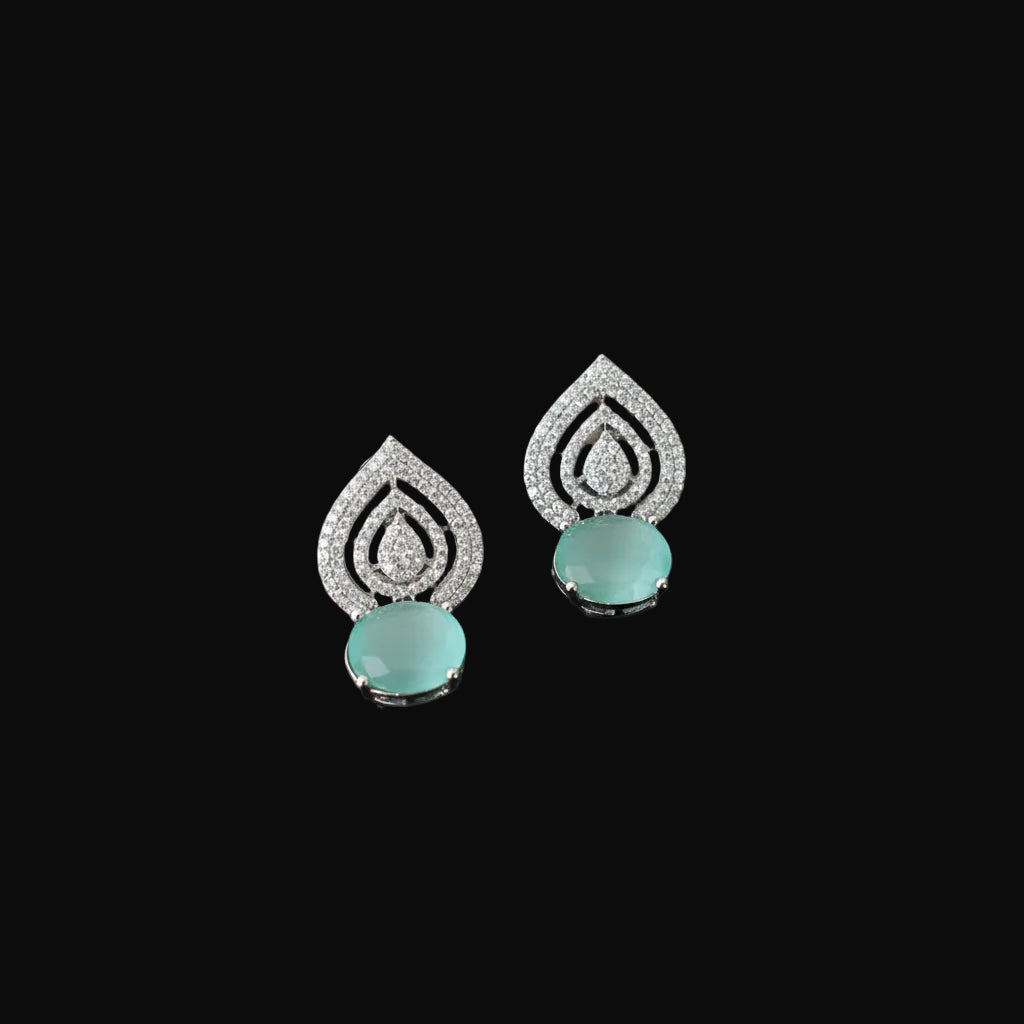 A pair of silver teardrop-shaped earrings with pavé-set clear stones and oval light green gemstone drops, displayed on a dark reflective surface.