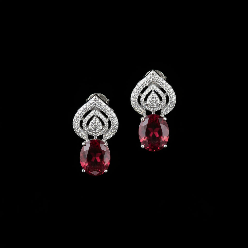 A pair of teardrop-shaped earrings with silver-toned pavé settings and oval red gemstones, displayed on a dark reflective surface.
