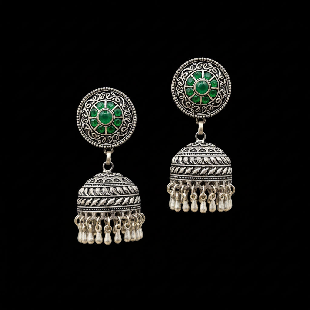 Ornate silver earrings with green stones on a gray stand, branded 'Saheli'.