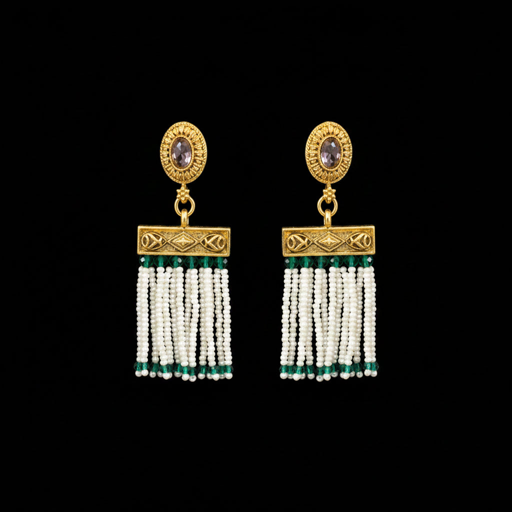 A pair of ornate gold-toned dangle earrings displayed on a dark gray Y-shaped jewelry stand. Each earring features a round gold top with a small oval stone, a rectangular gold connector, and multiple strands of white beads with small green accents.