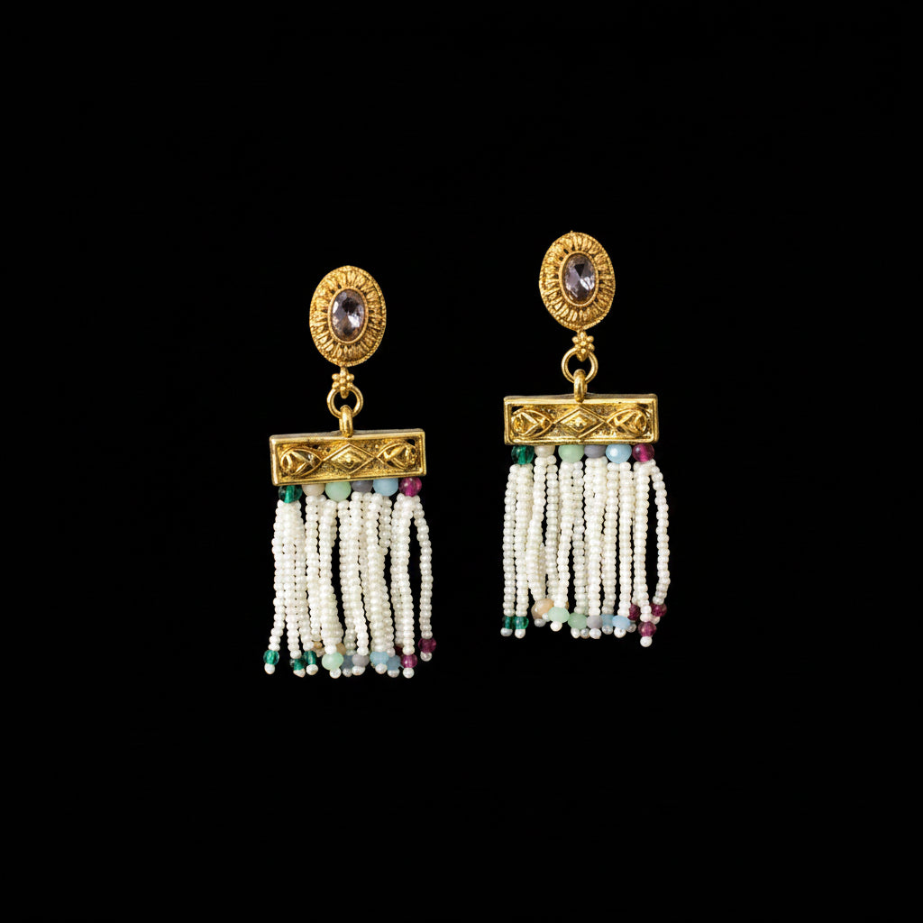 A pair of gold-toned traditional Indian dangle earrings with white beaded tassels and small multicolor bead accents, displayed on a black jewelry stand against a dark gray background.