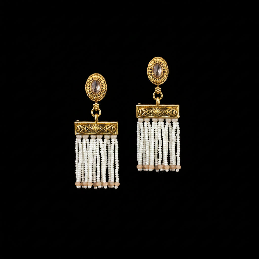 A pair of gold-toned statement earrings with oval stone studs and dangling beaded fringe, displayed on a dark gray Y-shaped jewelry stand against a dark gray background.