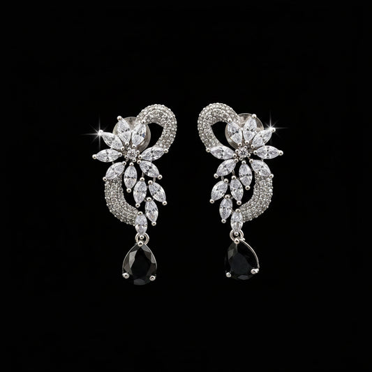 A pair of silver-tone drop earrings with clear marquise and round stones forming a floral heart design, each ending in a black teardrop stone, displayed on a black stand against a gray background. The brand logo 'SAHELI MY BEST FRIEND' is visible in the top left corner.