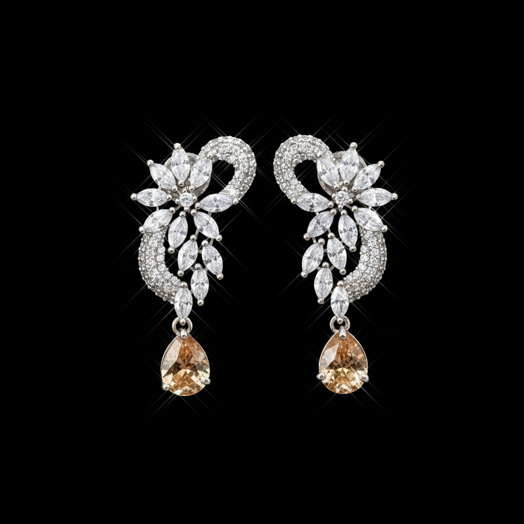 A pair of silver-toned statement earrings with clear marquise and round-cut stones, each featuring a dangling amber-orange teardrop gemstone, displayed on a black jewelry stand against a gray background. The brand logo 'SAHELI MY BEST FRIEND' is visible in the top left corner.