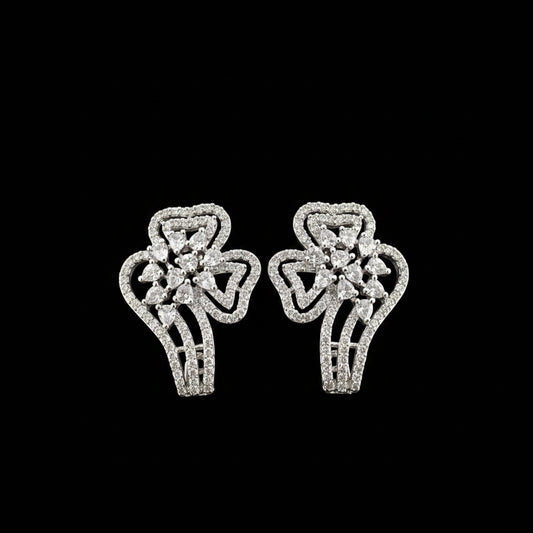A pair of silver-toned statement earrings with intricate pavé detailing, displayed on a black stand against a dark gray background with the brand logo 'SAHeli' at the top.