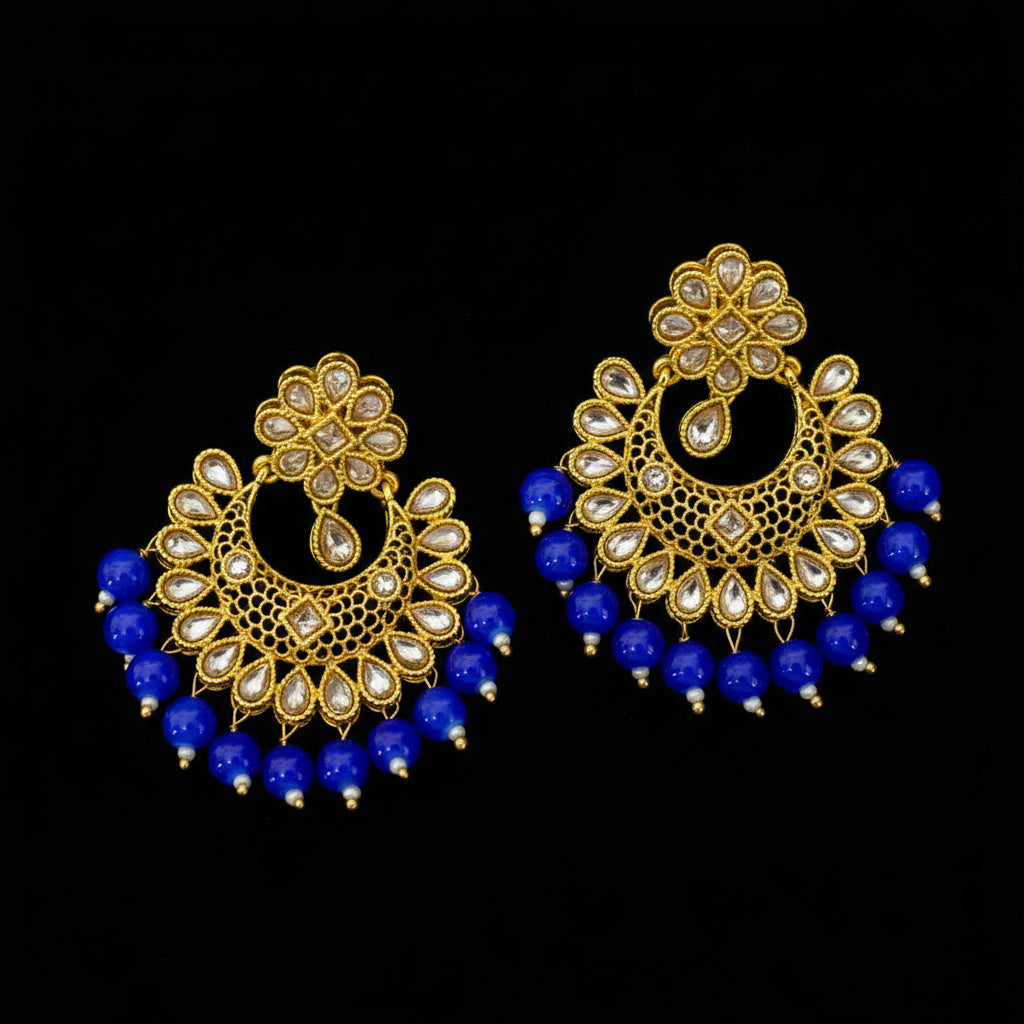 A pair of ornate gold-toned traditional Indian earrings with clear stone embellishments and dangling royal blue bead fringes, displayed on a dark gray surface. The brand logo 'SAHeli' is visible in the top left corner.
