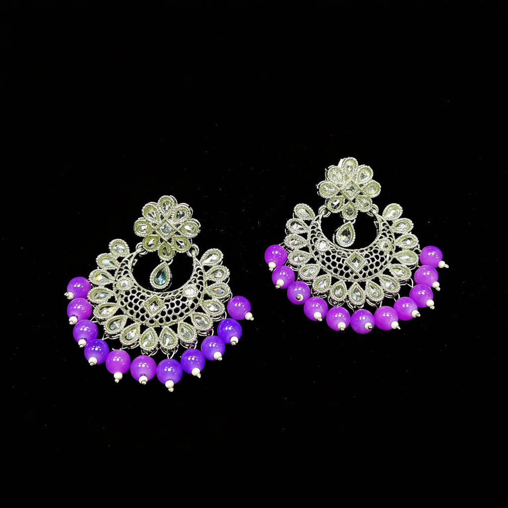 A pair of traditional Indian Jhumka earrings with silver-toned filigree work, clear rhinestones, and dangling purple beads, displayed on a black background with the brand logo 'SAHELI MY BEST FRIEND' above them.