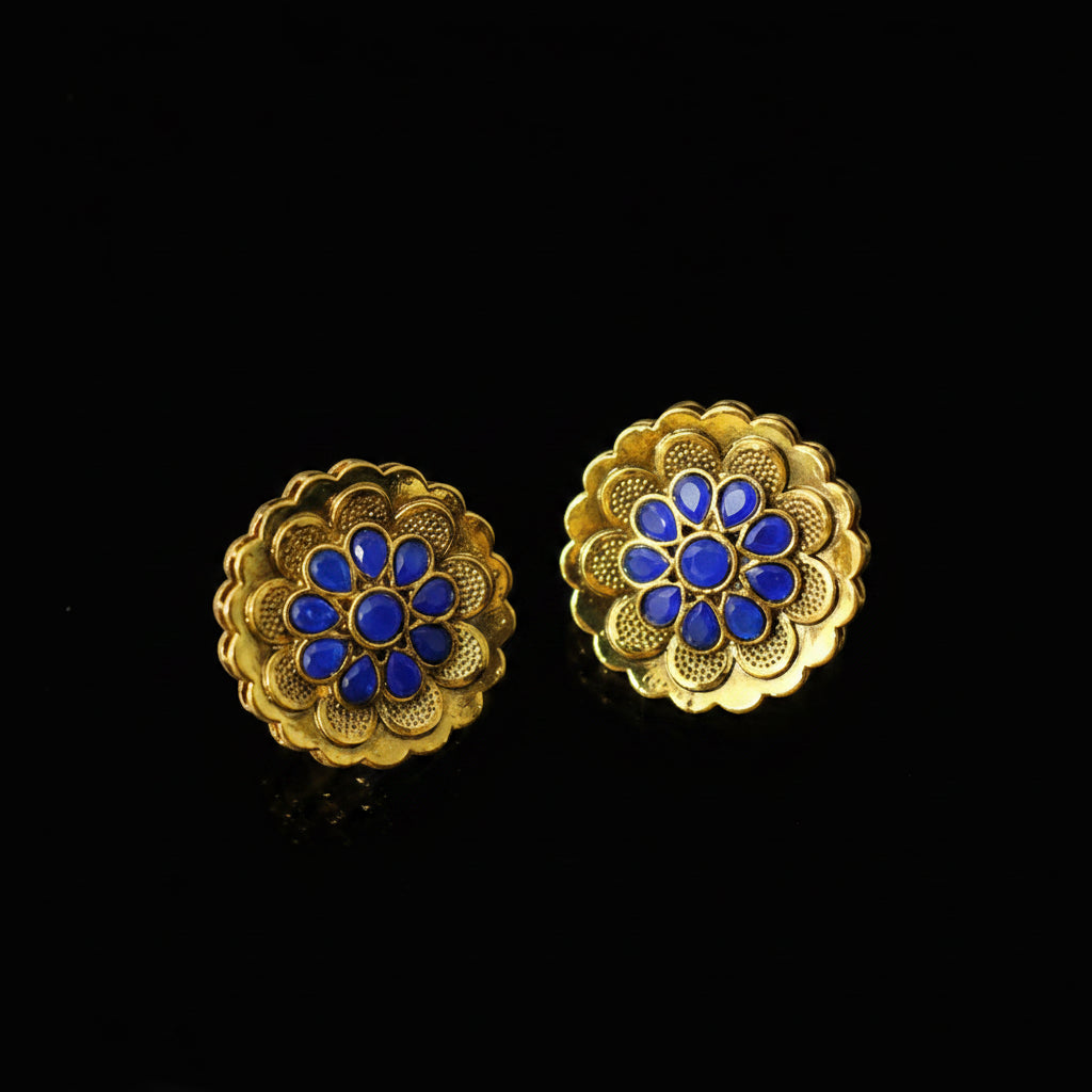 Gold floral earrings with blue centers on a dark background, featuring the brand 'Saheili'.