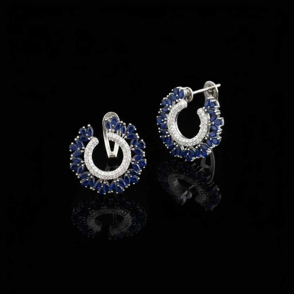 A pair of crescent-shaped hoop earrings with deep blue marquise-cut stones and a silver-toned center paved with small clear stones, displayed on a dark reflective surface with the SAHeli logo above.