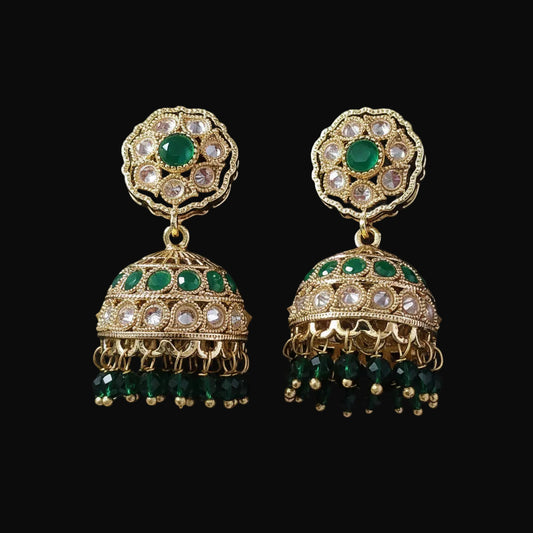 A pair of gold-plated Indian jhumka earrings with green stones and clear rhinestones, displayed on a solid dark pink background.