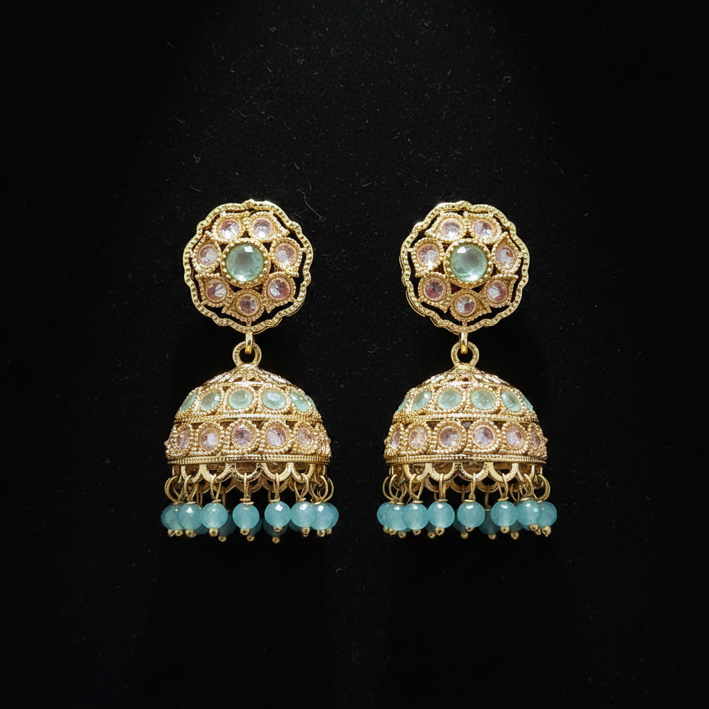 A pair of ornate gold-plated jhumka earrings with green stone accents and light blue bead drops, displayed on a solid dark pink background.