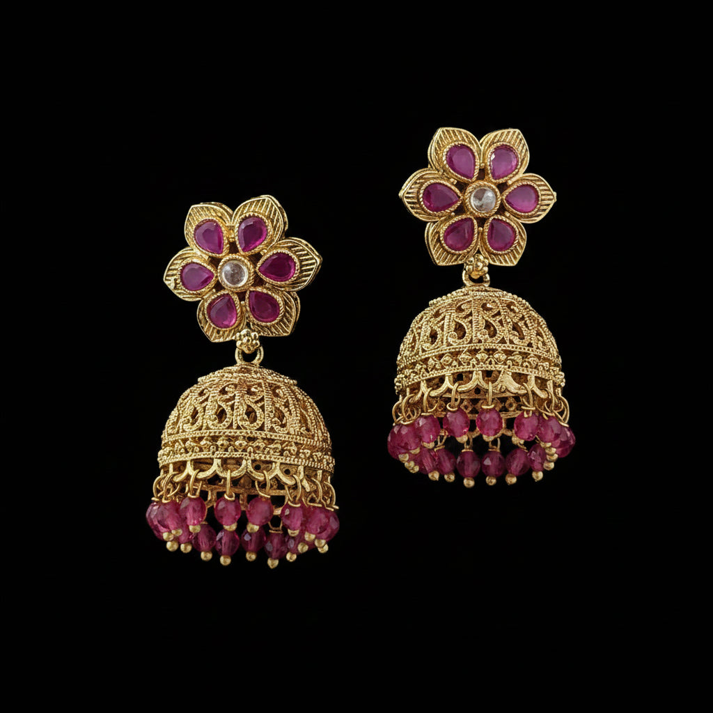A pair of traditional Indian gold-plated jhumka earrings with magenta pink gemstone accents and a central clear stone, displayed on a solid dark purple background.