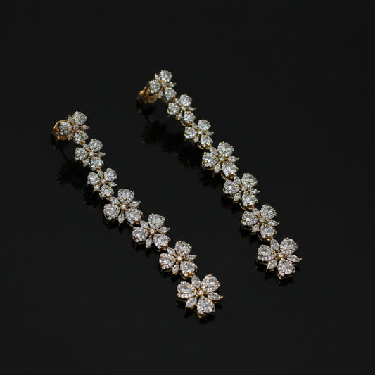 A pair of gold-toned drop earrings with a continuous row of flower-shaped clusters of clear stones, displayed on a dark gray background.