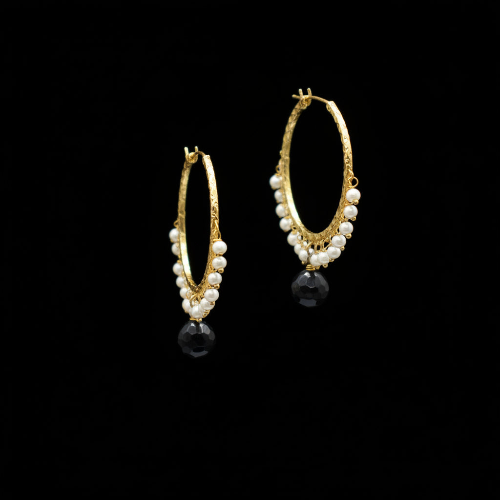 A pair of gold hoop earrings decorated with dangling white faux pearls and a large faceted black bead at the bottom, displayed on a dark gray background.