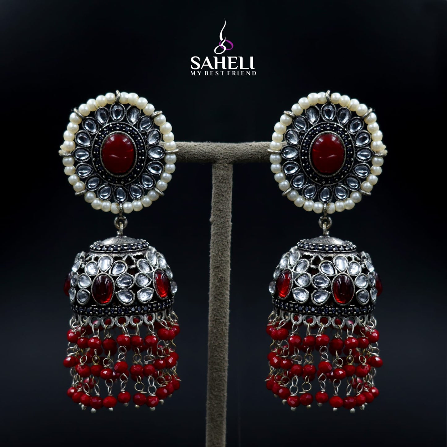 Pair of ornate earrings with red and silver details on a dark background, featuring the brand 'Saheli'.