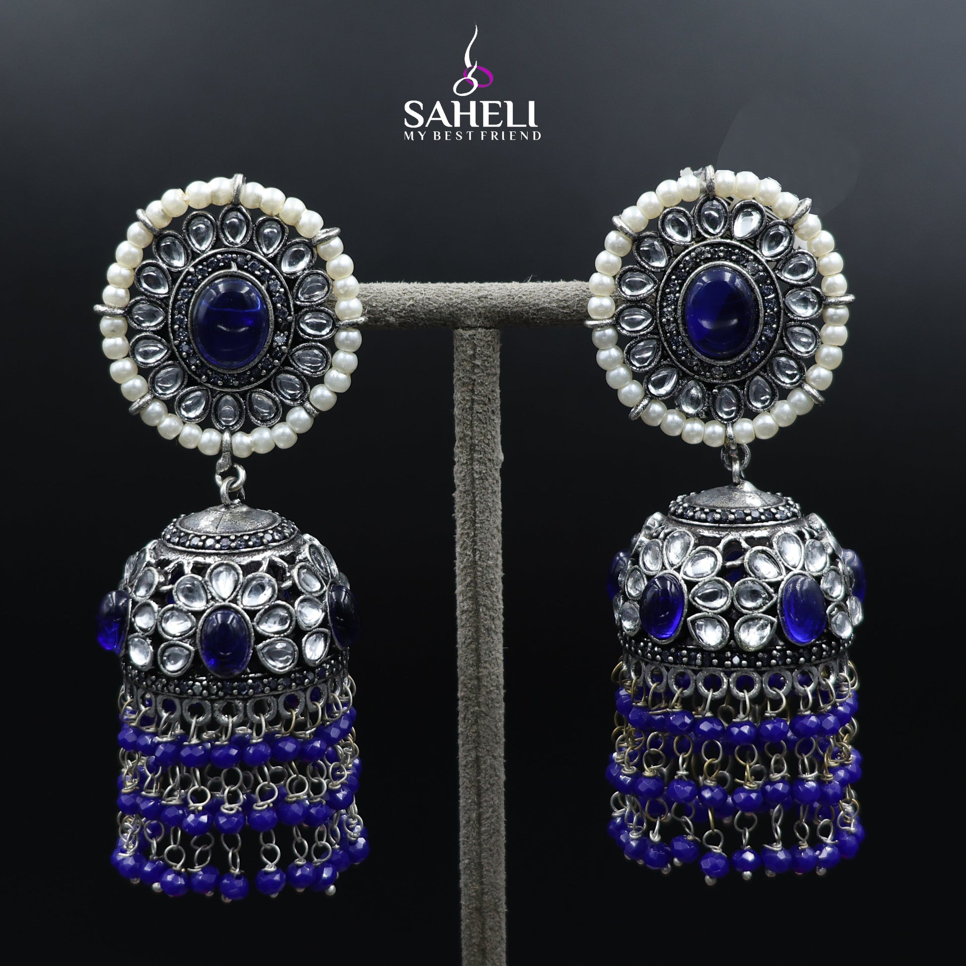 Pair of earrings with blue stones and pearl-like beads on a dark background, branded 'Saheli'.