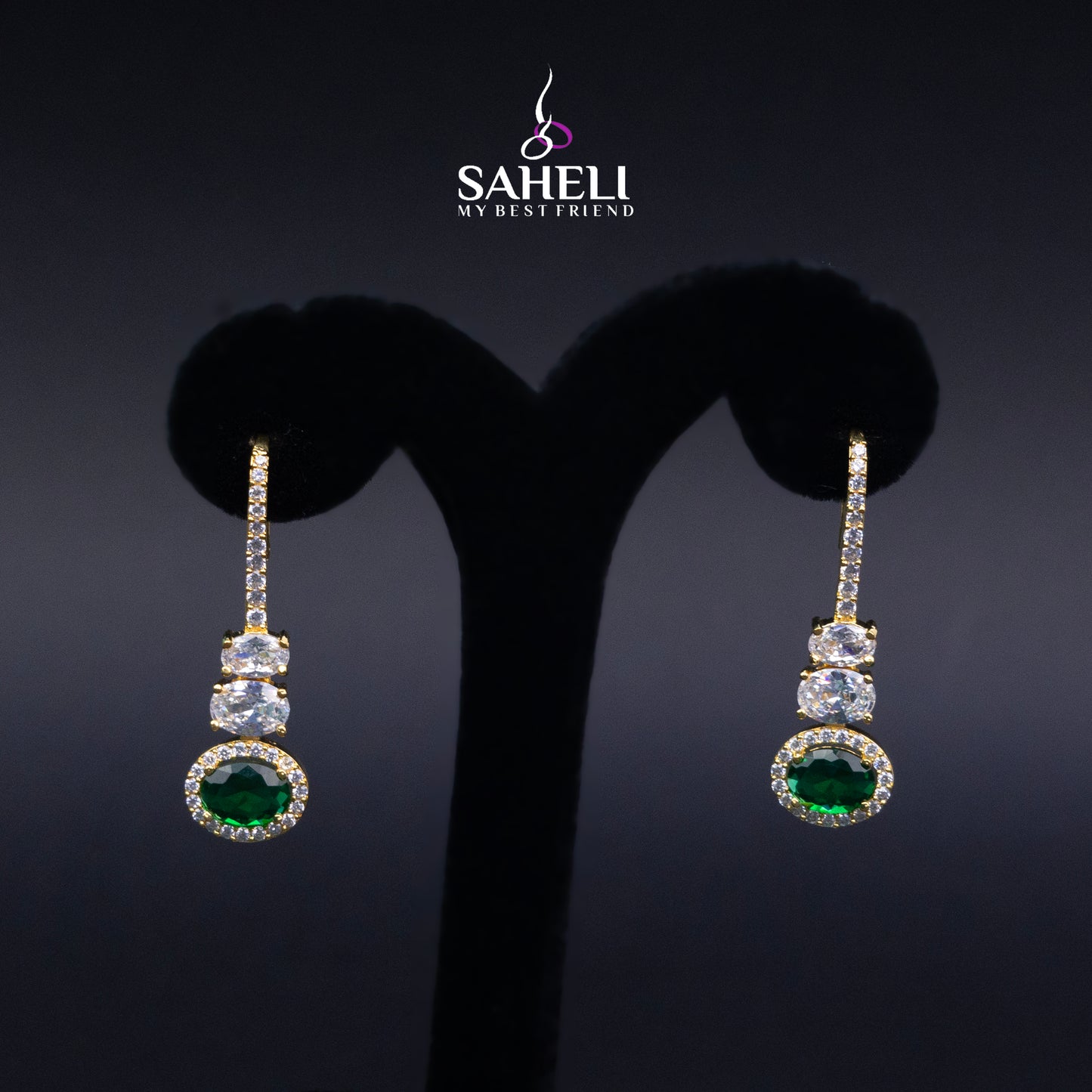 Pair of earrings with green gemstones on a black background, featuring the brand 'Saheli'.