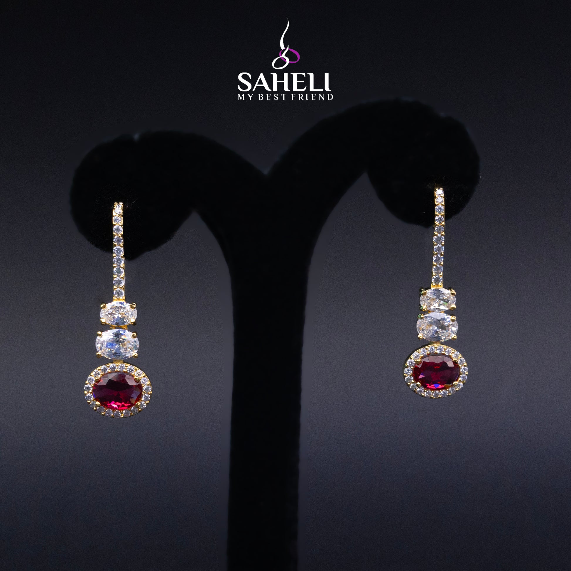Pair of earrings with gemstones on a black stand, branded 'Saheli'.