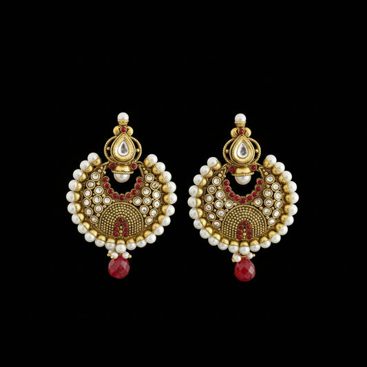 A pair of traditional Indian gold-plated earrings decorated with white pearls, red stones, and a central black bead, displayed on a dark gray surface with the SAHELI brand logo above.
