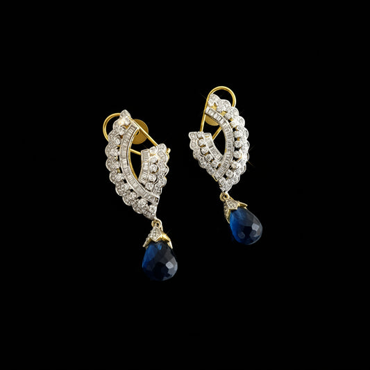 A pair of gold and silver drop earrings with clear pavé stones and deep blue teardrop crystals, displayed on a reflective gray surface with the SAHELI brand logo above.