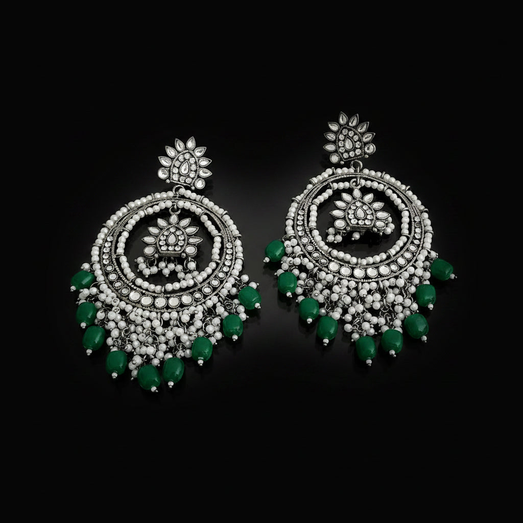 A pair of silver-toned Indian chandelier earrings decorated with white beads, clear stones, and green bead accents, displayed on a dark reflective surface.
