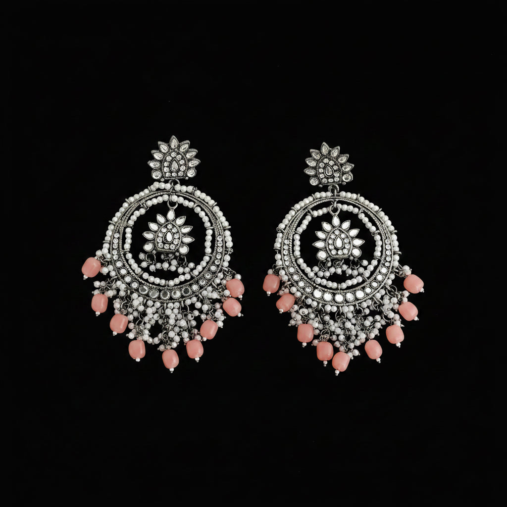A pair of ornate silver-toned dangle earrings decorated with white stones and pink bead tassels, displayed on a dark reflective surface.