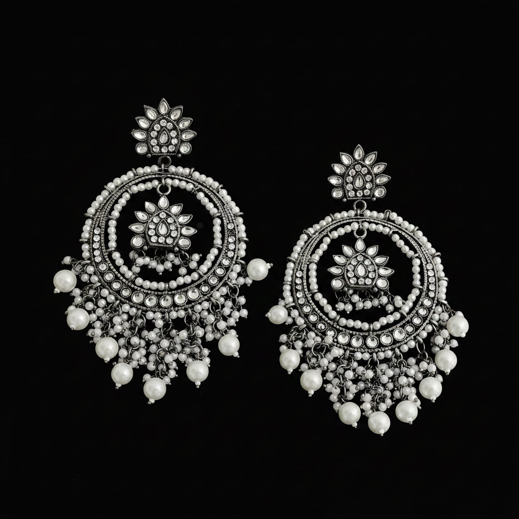 A pair of ornate silver-toned dangle earrings with intricate circular beadwork, floral motifs, and dangling pearl-like beads, displayed on a dark reflective surface.