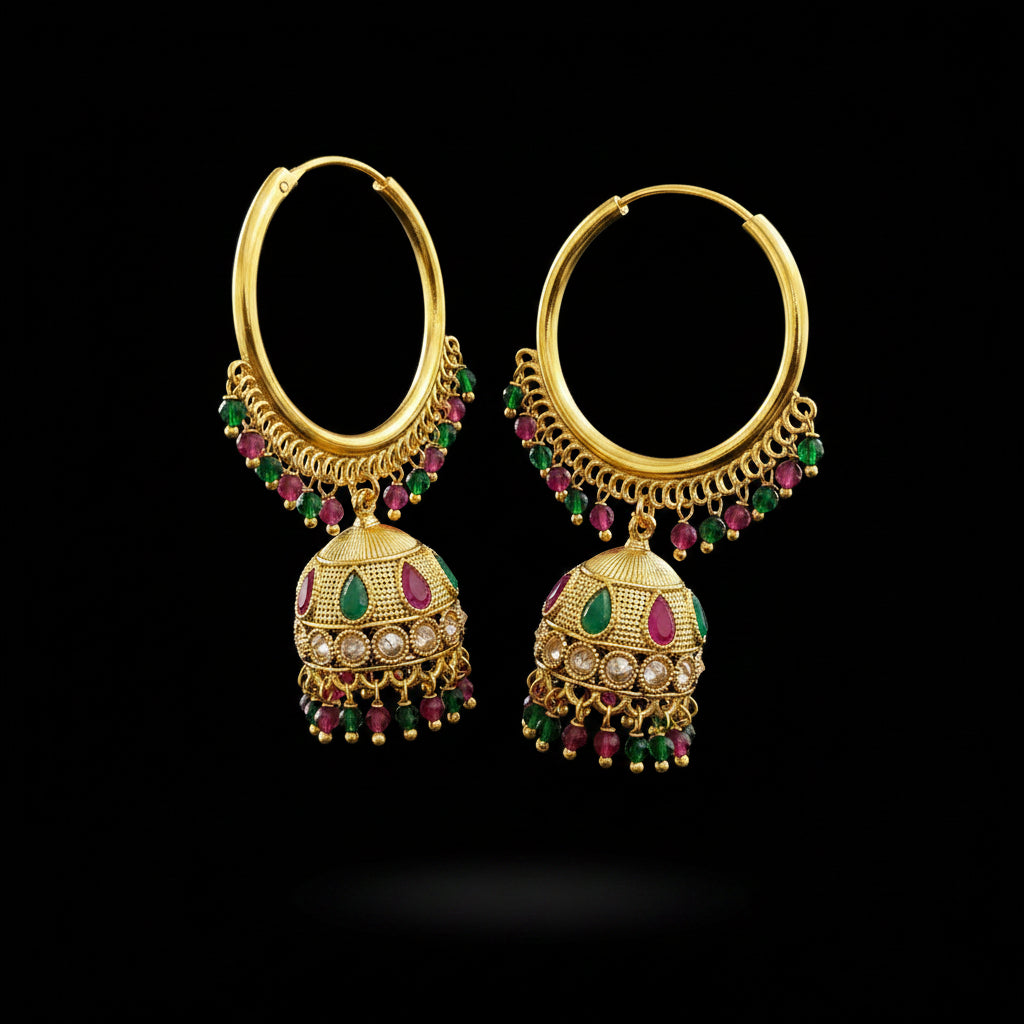 A pair of gold-plated Indian jhumka earrings with green and pink gemstone accents, displayed on a gray stand against a light gray background.