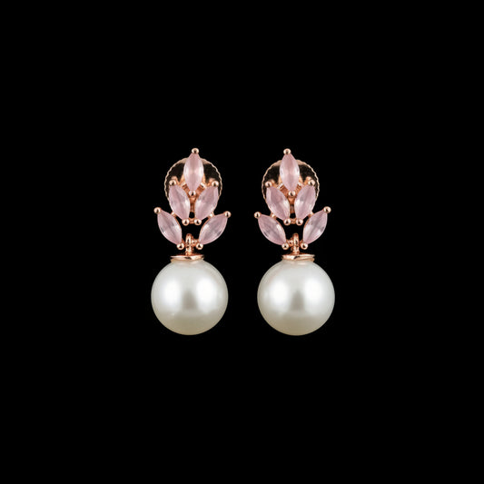 A pair of rose gold floral stud earrings with pale pink stones and a white pearl drop, displayed on a black textured stand against a dark gray background.