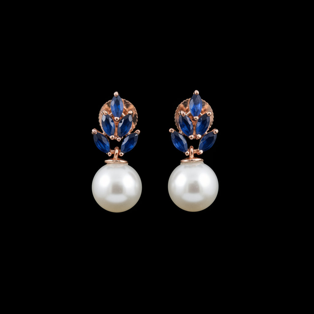 A pair of rose gold and blue gemstone earrings with large white pearl drops, displayed on a black stand against a dark gray background.