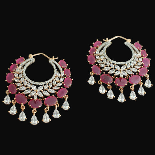 A pair of gold-plated hoop earrings decorated with pinkish-red round gemstones, clear pavé-style stones, and dangling clear teardrop-shaped crystals, displayed on a dark reflective surface.