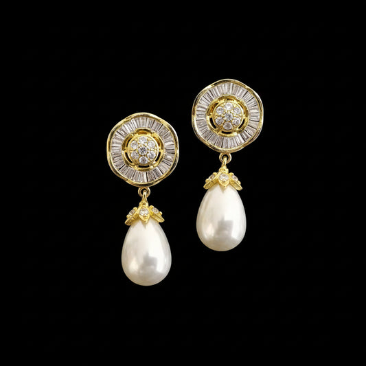 A pair of gold and white pearl drop earrings displayed on a black jewelry stand against a gray background, with the SAHELI by HEARTBEAT logo printed above.