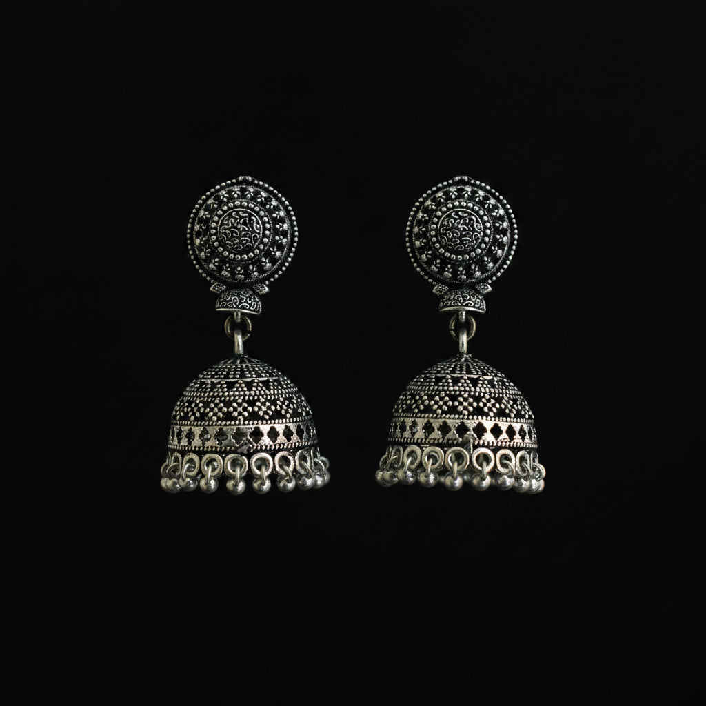 A pair of ornate silver-toned jhumka earrings with intricate filigree detailing, displayed on a decorative green and blue feather against a dark background.