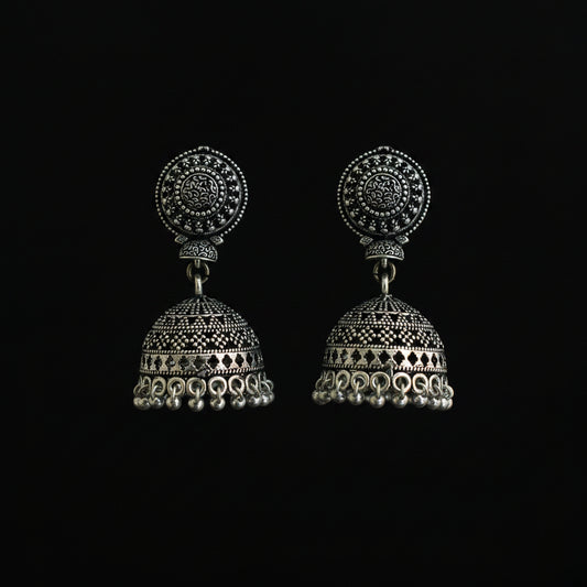 A pair of ornate silver-toned jhumka earrings with intricate filigree detailing, displayed on a decorative green and blue feather against a dark background.