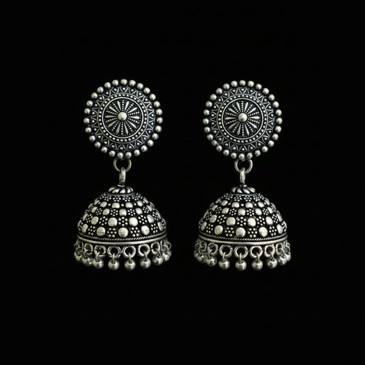 A pair of silver-toned Indian jhumka earrings with intricate filigree and beadwork, displayed on a green and blue peacock feather against a dark reflective background.