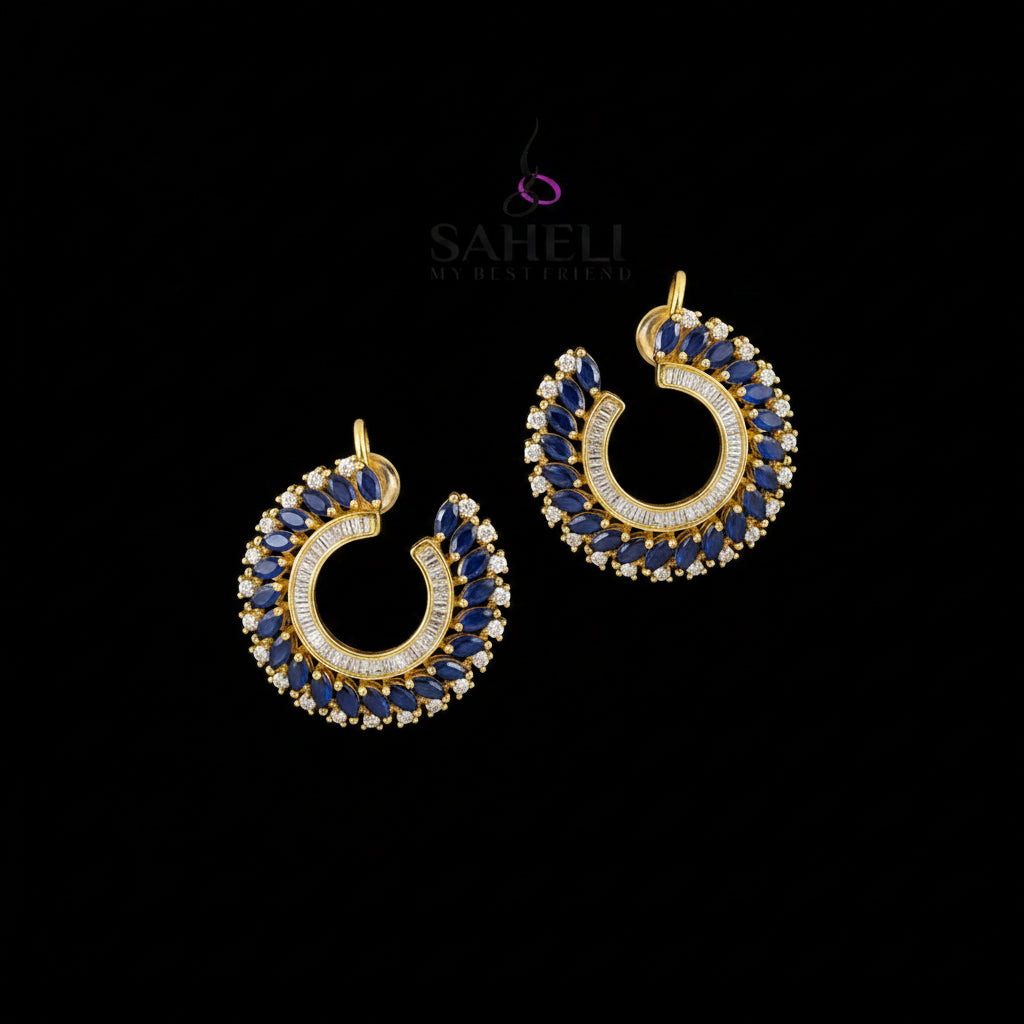 A pair of gold crescent-shaped hoop earrings decorated with blue and clear stones, displayed on a dark reflective surface with the SAHELI brand logo above them.