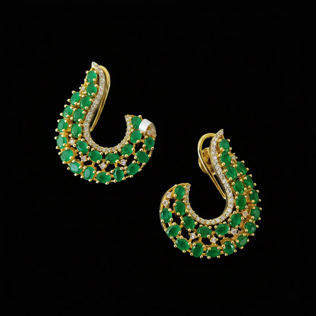 A pair of gold crescent-shaped hoop earrings embellished with green stones and small clear accent stones, displayed on a dark reflective surface with the SAHeli My Best Friend logo in the background.