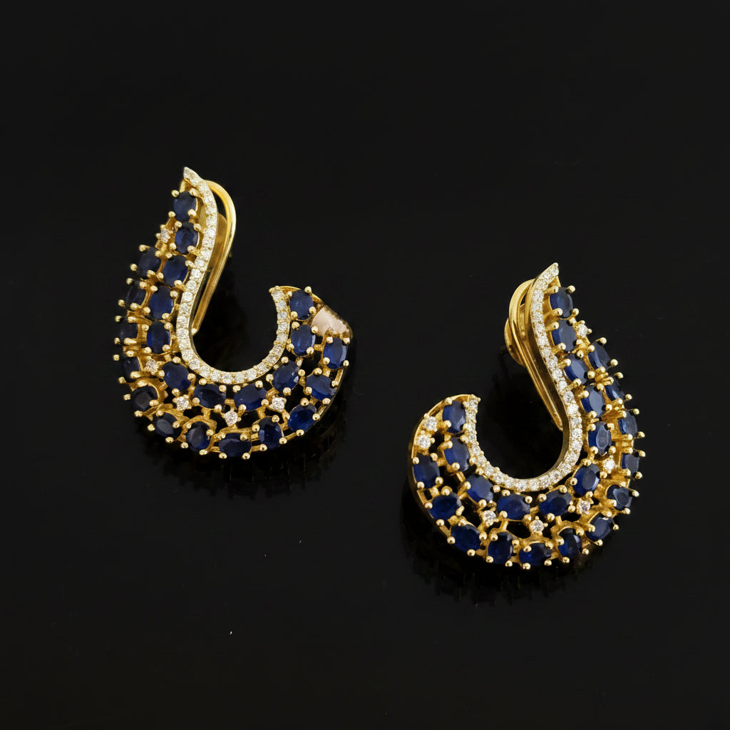 A pair of gold crescent-shaped stud earrings embellished with deep blue round stones and small clear accent stones, displayed on a dark reflective surface with the SAHELL MY BEST FRIEND logo above.
