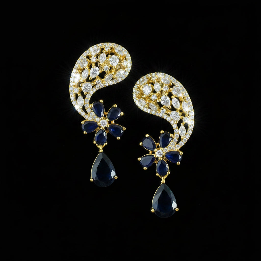 A pair of gold-toned traditional Indian jhumka earrings with deep blue stones and clear pavé accents, displayed on a dark reflective surface with a Saheli by Srinivasand logo in the top left corner.