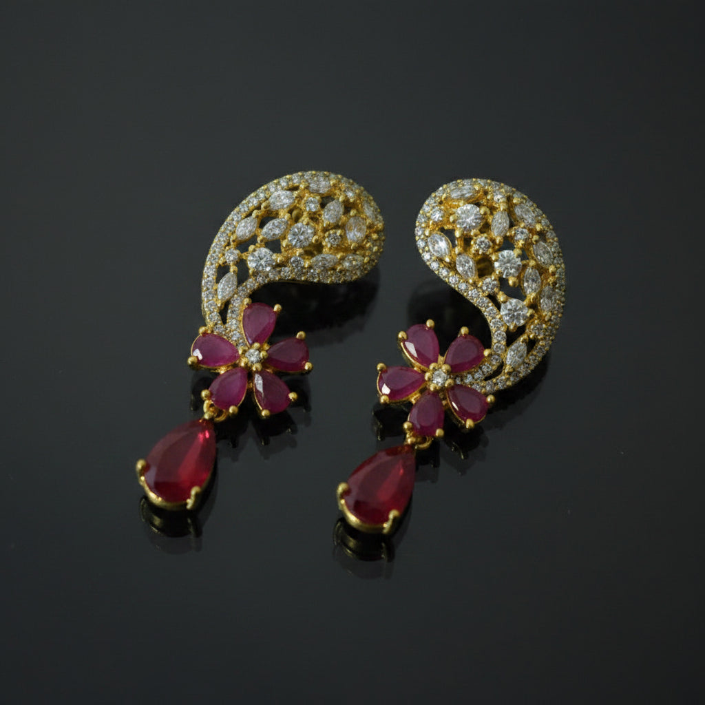 A pair of gold crescent-shaped earrings decorated with clear crystals and red teardrop gemstones, displayed on a black reflective surface with a SAHELI MY BEST FRIEND logo in the top right.